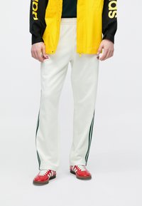 Λευκό παντελόνι jogging με πράσινες λωρίδες στο πλάι, φορεμένο με φωτεινό κίτρινο μπουφάν και κόκκινα αθλητικά παπούτσια με άσπρα κορδόνια.