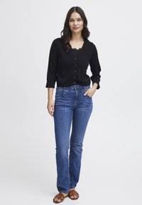Svart knapp-up cardigan med V-ringning, tillsammans med blå utsvängda jeans. Modellen bär bruna sandaletter med remmar. Enkel, klassisk design.