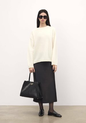 Maglione crema, gonna nera, occhiali da sole neri. Borsa in pelle nera con motivo coccodrillo e placchetta. Mocassini neri. Sfondo a pavimento liscio.
