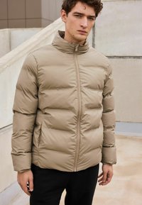 Jachetă puffer bej cu guler înalt, având cusături orizontale și fermoar frontal. Asortată cu pantaloni negri pentru contrast.