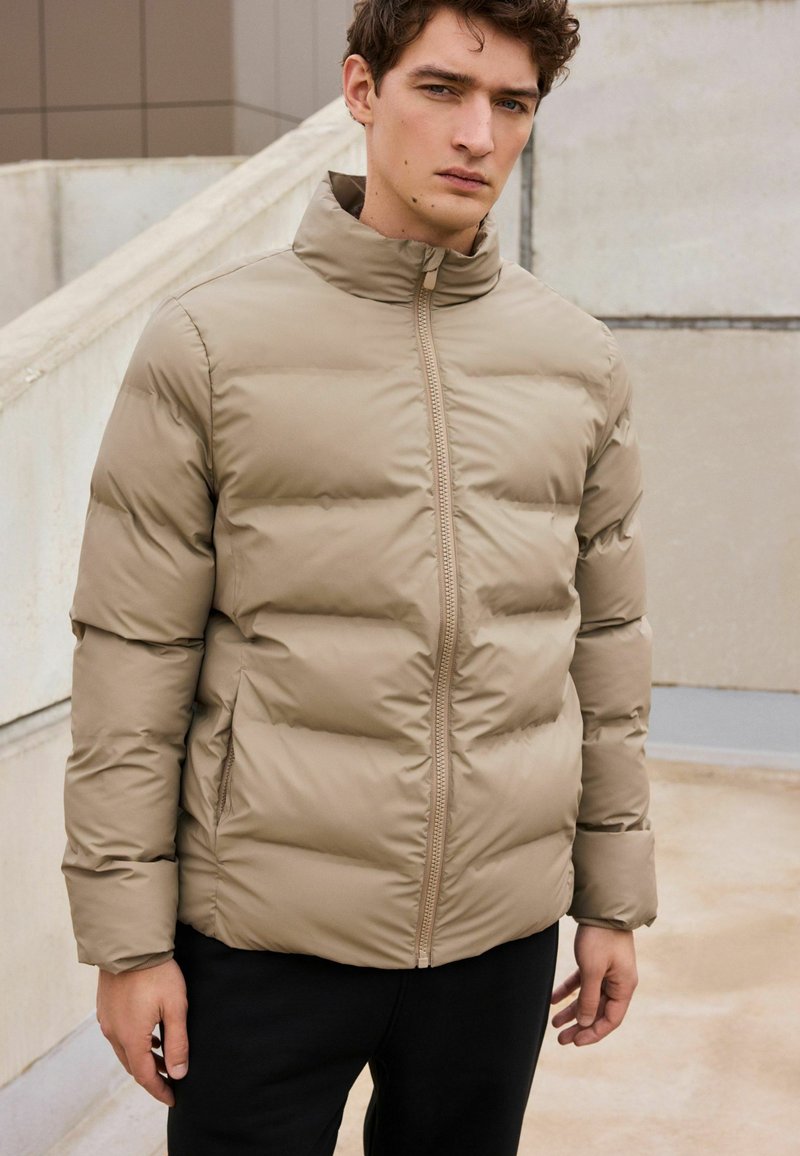 Jachetă puffer bej cu guler înalt, având cusături orizontale și fermoar frontal. Asortată cu pantaloni negri pentru contrast.