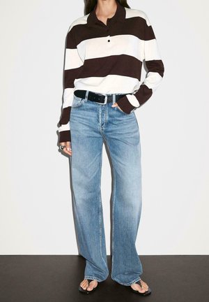 Osoba ubrana w brązowo-białą koszulkę polo z długim rękawem w paski, jasnoniebieskie szerokie dżinsy, czarny pasek i czarne klapki, stojąca przy białej ścianie.