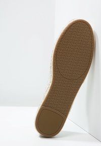 Zapatilla texturizada en beige con suela de goma, que presenta un dibujo en la superficie y una pestaña trasera para tirar. El material superior es tejido y ligero.