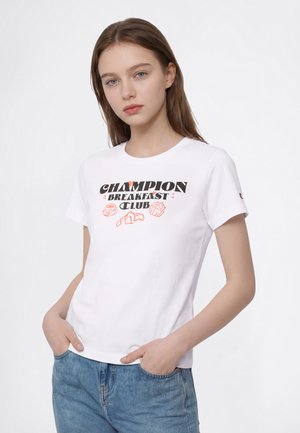 Млада жена с дълга кафява коса, носеща бяла тениска с текст "Champion Breakfast Club" и икони за закуска, комбинирана със сини дънки.