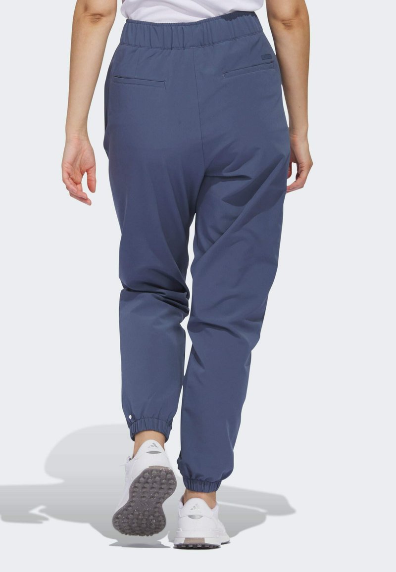Personne marchant loin, portant un pantalon de jogging élastique bleu et des baskets blanches à semelles texturées sur un fond clair.