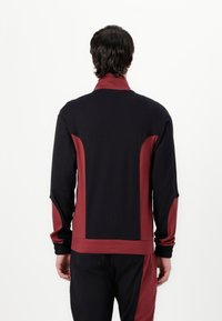 Giacca sportiva nera e bordeaux con colletto alto, pannelli laterali a contrasto e polsini elastici. Tessuto morbido e design dal taglio rilassato.