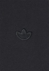 Svart ribbad textil med en upphöjd svart broderad trefoil-logotyp i mitten. Bakgrunden visar en jämn ribbad textur.