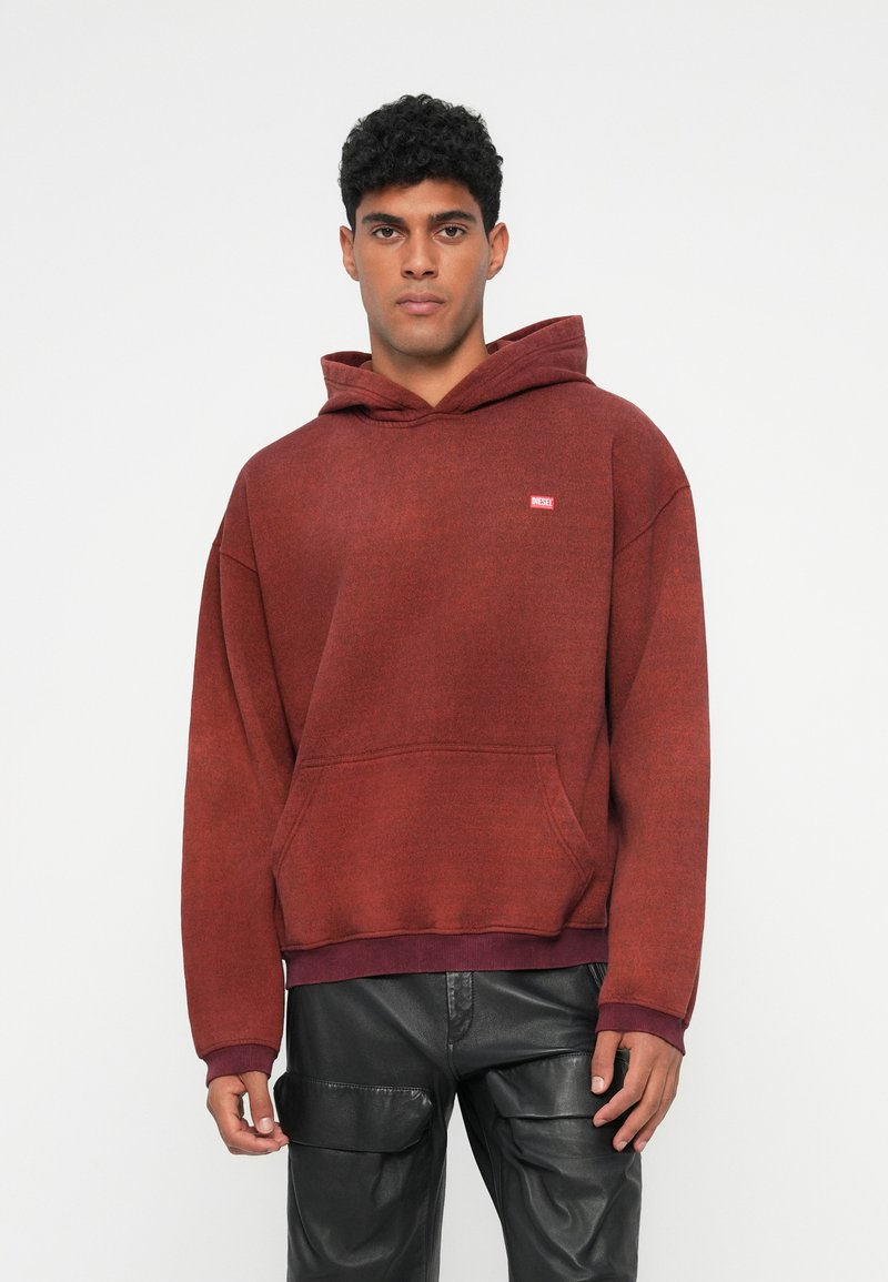 Sudadera burdeos hecha de tejido texturizado, con bolsillo frontal, puños acanalados y un logo bordado. Combinada con pantalones cargo negros.