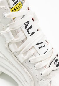 Baskets montantes en cuir blanc avec des lacets blancs épais, des étiquettes de marque noires et jaunes, et des œillets en métal argenté ; détail de surface texturée visible.