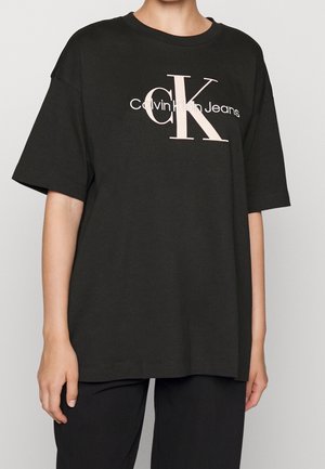 Osoba nosící volné černé tričko s nápisem "Calvin Klein Jeans" a velkým logem "cK" v světle béžové barvě na přední straně, doplněné černými kalhotami.