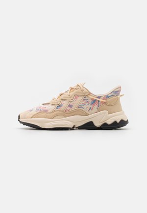 Beżowy sneaker z wielokolorowym abstrakcyjnym wzorem, falistym wzorem podeszwy, teksturowanymi nakładkami ze skóry oraz czarną podeszwą na jednolitym białym tle.