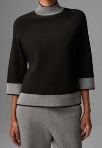 Svart topp med hög ribbad krage, trekvartsärmar och grå randiga detaljer vid ärmar och nederkant. Mjuk och slät material.