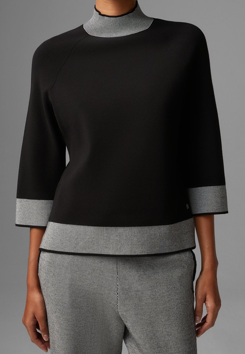Svart topp med hög ribbad krage, trekvartsärmar och grå randiga detaljer vid ärmar och nederkant. Mjuk och slät material.