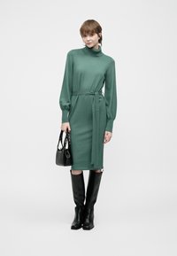 Grønt strikket kjole med høy hals, lange ermer og belte i midjen. Kombinert med svarte over kne støvler og en liten svart veske.