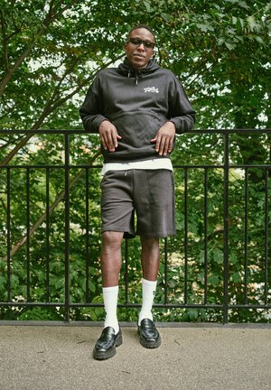 Mann trägt schwarzen Kapuzenpullover, schwarze Shorts, weiße Socken und schwarze Schuhe und lehnt an einem schwarzen Metallgeländer, im Hintergrund grüne, belaubte Bäume.