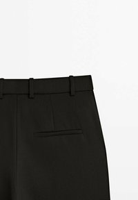 Pantalon noir ajusté en tissu texturé. Les caractéristiques incluent un devant plat, des passants de ceinture et une poche arrière unique.