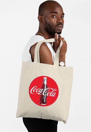 Beżowa torba bawełniana z czerwoną okrągłą naszywką z ilustracją butelki Coca-Cola w stylu vintage oraz logo w białym skrypcie, mocne uchwyty.