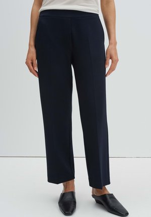 Person, der schwarze gerade geschnittene Hosen und schwarze Slipper trägt, steht vor einem schlichten Hintergrund.