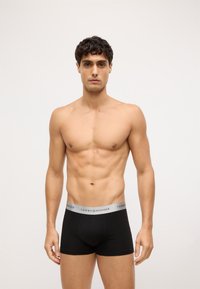 Boxer shorts negros de algodón con una cinturilla elástica gris que presenta el logo de "TOMMY HILFIGER". El modelo está de pie frente a un fondo neutro.