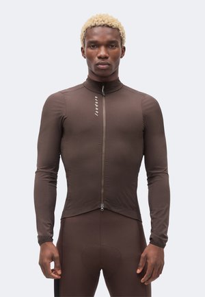 Mann mit kurzen lockigen blonden Haaren trägt eng anliegendes braunes langärmeliges Radtrikot und passende Shorts vor einfarbigem Hintergrund.