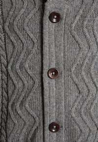 Cárdigan gris de punto que presenta un patrón texturizado en zigzag, botones marrones y acanalado vertical a lo largo de la tapeta de los botones.