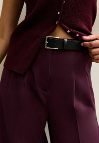 Bordeaux breiten gebreide cardigan met gouden knopen, gecombineerd met hoge, op maat gemaakte broeken in een bijpassende tint en een zwarte leren riem met een gouden gesp.