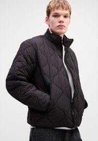 Young man wearing black quilted zip-up jacket with hands in pockets, white shirt underneath, and gray pants against plain background.