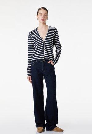 Vrouw staat met hand in zak, draagt een marineblauw-wit gestreepte knoopvest, donkere wijde jeans en beige loafers.