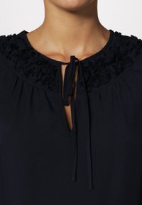 Námořnická modrá blůza s volánkovým límcem a zavazováním u krku, s malým výstřihem a měkkou texturou látky.