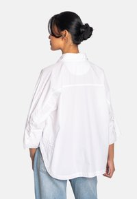 Hvit skjorte uten krage med trekvart ermer, som har knappdetaljer og en avslappet passform. Stoffet virker lett og har en glatt tekstur.