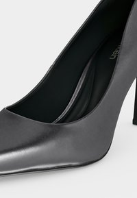 Chaussure à talon haut en cuir noir à bout pointu avec talon stiletto fin et finition lisse sur fond blanc.