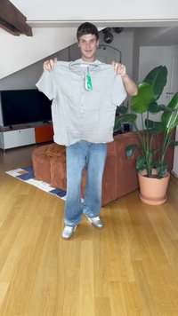 Camicia grigia chiaro a maniche corte con bottoni tenuta da una persona, con un'etichetta. Sullo sfondo ci sono un divano marrone e una pianta in vaso.