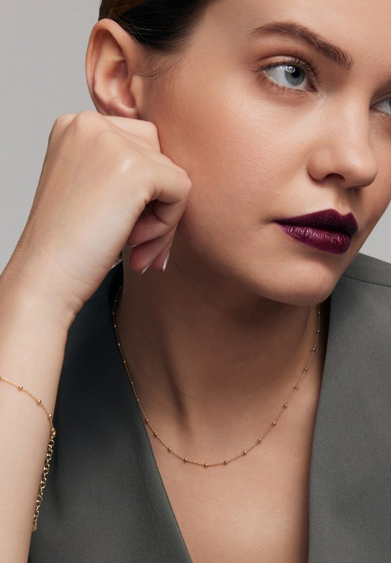 Złoty naszyjnik z małymi, równomiernie rozmieszczonymi okrągłymi koralikami, noszony z szarym odzieżą. Skóra jest gładka; ciemna szminka podkreśla wygląd.