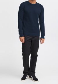 Marineblauer Strickpullover mit strukturiertem Muster, rundem Halsausschnitt und gerippten Bündchen, kombiniert mit schwarzen Hosen und schwarzen Sneakern mit weißen Akzenten.