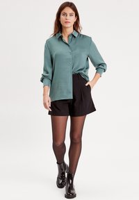 Blouse en soie teal avec des boutons, associée à un short noir et des collants opaques. Des bottines noires brillent et complètent la tenue décontractée.