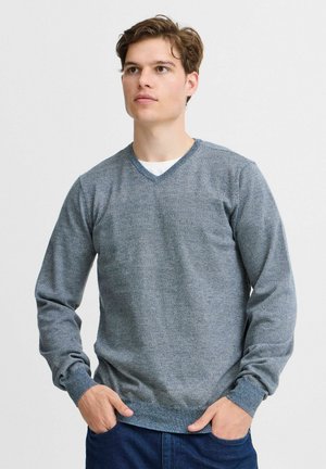 Jeune homme aux cheveux bruns courts portant un pull bleu en V et un jean bleu foncé, les mains dans les poches, regardant légèrement vers le haut.