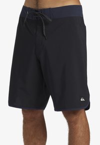 Shorts de bain noirs pour homme avec un design uni, dotés d'une ceinture bleu marine, d'un cordon de serrage, d'un tissu texturé et d'un petit logo sur la jambe gauche.