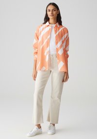 Oranje overhemd met witte diagonale strepen, gecombineerd met crèmekleurige uitlopende broek en witte sneakers. Soepele stof, ontspannen pasvorm.