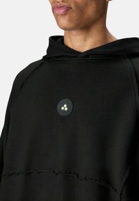 Zwarte hoodie met een ronde, zwarte accentkleur en drie lichtgekleurde stippen. Gemaakt van zachte stof met een losse pasvorm en unieke stiksel details.