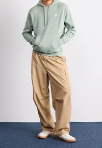 Persona con sudadera Nike verde claro, pantalones cargo beige y zapatillas blancas, de pie sobre una alfombra azul contra una pared blanca.