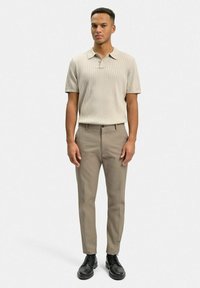 Beige ribbad kortärmad pikétröja och ljusbruna skräddarsydda byxor, tillsammans med svarta snörskor. Enfärgad bakgrund.