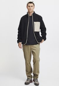 Black fleece jacket with beige pocket and trim, paired with khaki pants. Notable details include a zippered pocket and textured fabric.