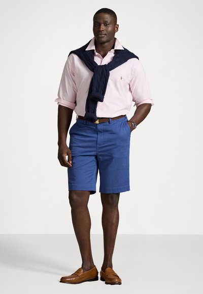 Suéter de algodão azul-marinho drapeado sobre uma camisa listrada rosa claro, combinado com calções azuis e mocassins castanhos. Relógio simples no pulso.