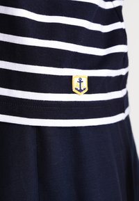 Abito blu navy con strisce orizzontali bianche. Presenta un distintivo di un'ancora giallo e blu in vita, realizzato in tessuto di cotone.