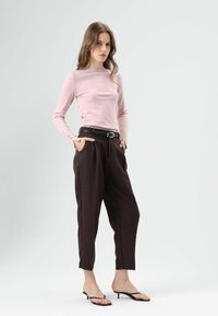 Lichtroze top met lange mouwen, strakke pasvorm en knoopdetails op de mouwen, gecombineerd met losse bruine broek en zwarte sandalen met hak.