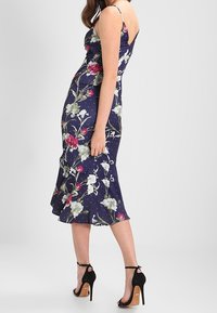Abito a stampa floreale con sfondo blu navy, corpetto aderente, spalline sottili e orlo svasato. Abbinato a sandali neri con tacco alto.