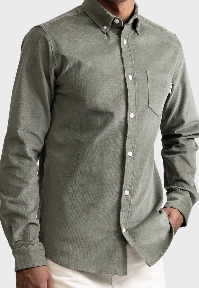 Homme portant une chemise en velours côtelé vert olive clair à manches longues avec une poche poitrine, main dans la poche, sur un fond uni.