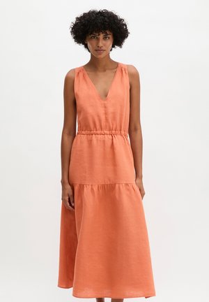 Frau mit lockigem Haar, die ein ärmelloses, V-Ausschnitt, locker sitzendes orangenes Midikleid mit geraffter Taille trägt, steht vor weißem Hintergrund.