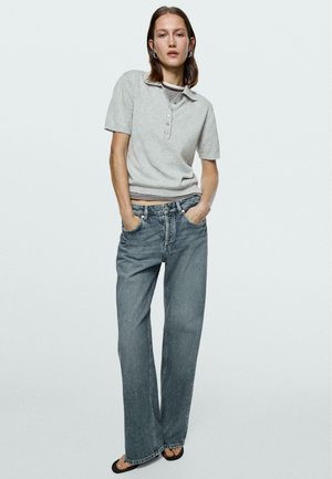 Kvinde iført en grå kortærmet trøje med krave over en stribet top, kombineret med løstsiddende, falmede blå jeans og sorte sandaler.