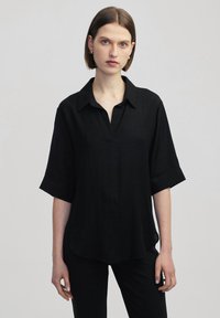 Blusa nera a maniche corte con colletto, caratterizzata da una vestibilità comoda e un tessuto leggermente texturizzato. Immortalata su uno sfondo chiaro.